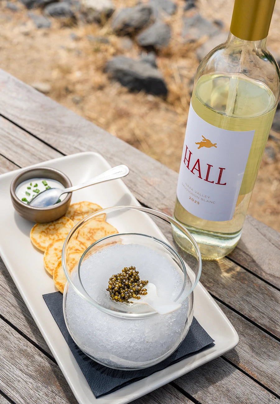 A bottle of Hall white wine sits on a wooden table next to a glass of iced white wine with a berry garnish, accompanied by crackers and a small dipping sauce on a white tray.
