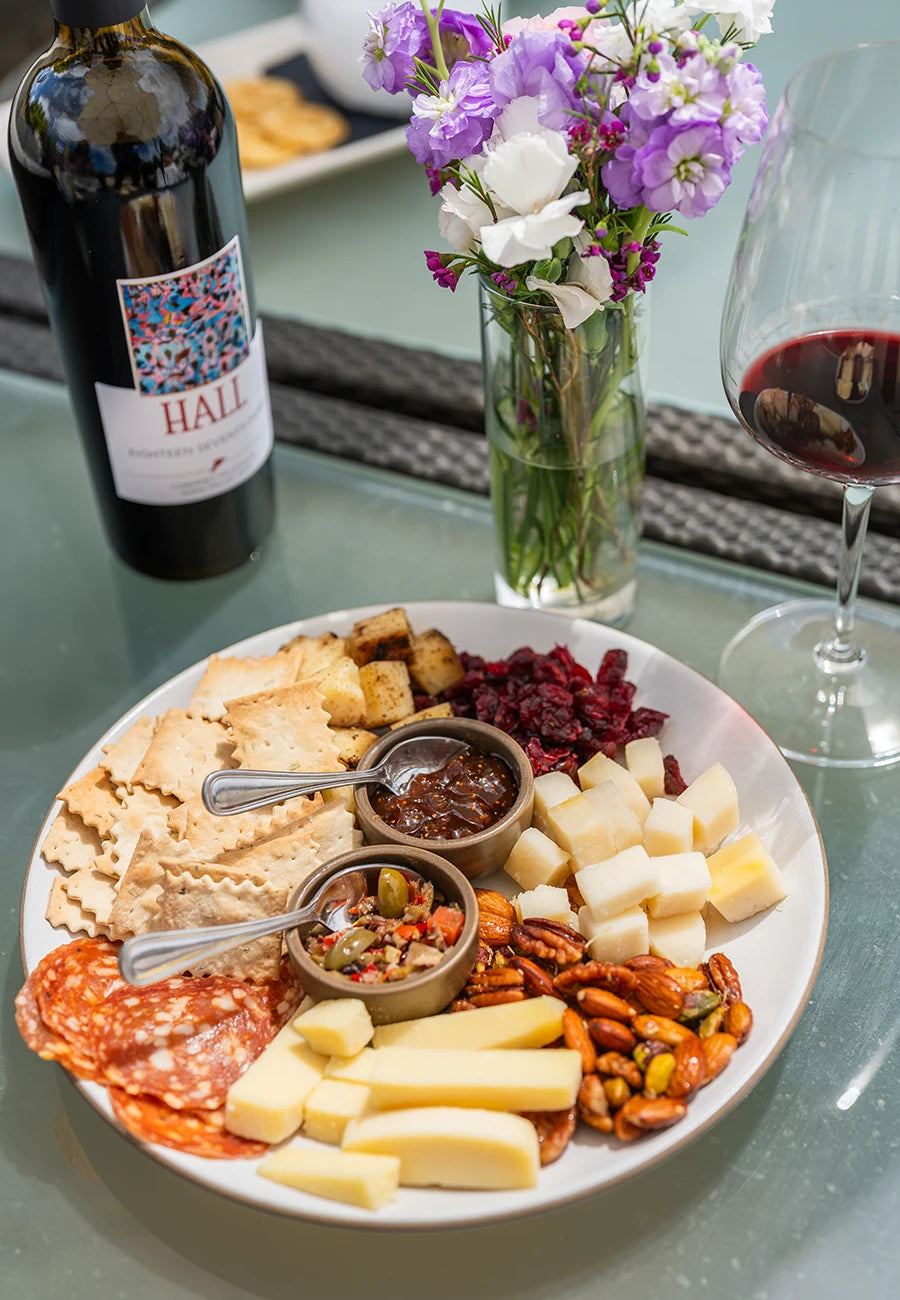 A charcuterie board with cheese, crackers, salami, nuts, dried fruit, and small bowls of dip on a glass table, alongside a bottle of Hall wine, a glass of red wine, and a vase of flowers.