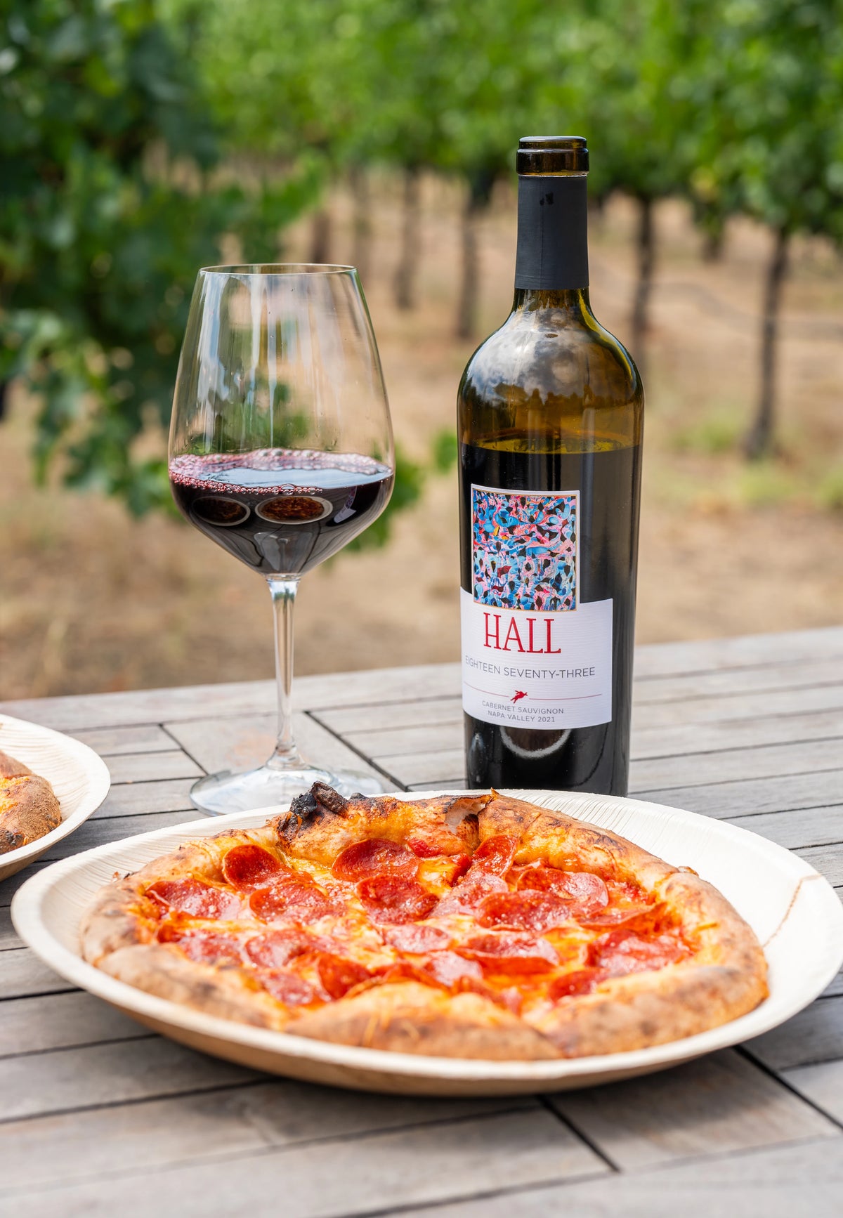 A bottle of Hall Non-Alcoholic Red Wine and a filled wine glass on a wooden table next to a pizza, with a blurred vineyard background.